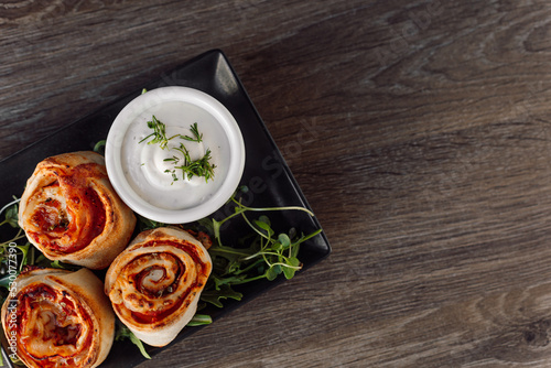 Appetizing pepperoni pastry rolls with filling, greenery and sauce on black elegant plate on wooden table in restaurant