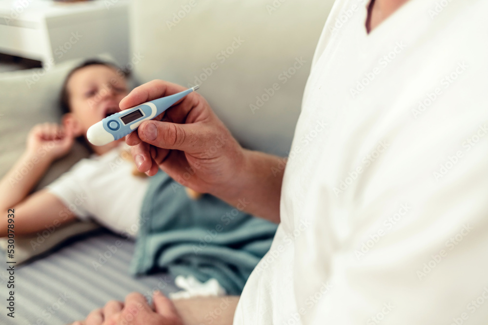 Photo of Sick male kid with high fever laying in bed while father taking temperature. Cropped shot of a man checking his son's temperature.