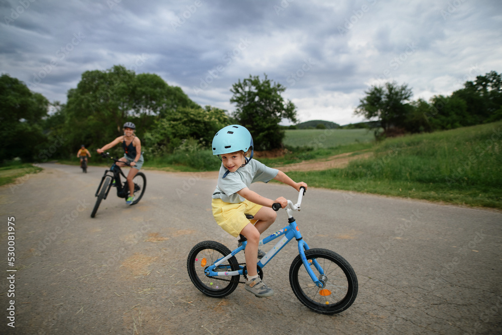 Portrait of excited little boy with his family at backround riding bike on path in park in summer Portrait of excited little boy with his family at backround riding bike on path in park in summer