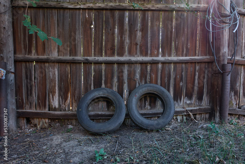 Two old wheels near the fence. Old car tires for a sedan are thrown away near an old wooden fence. Fence rotten covered with mold.