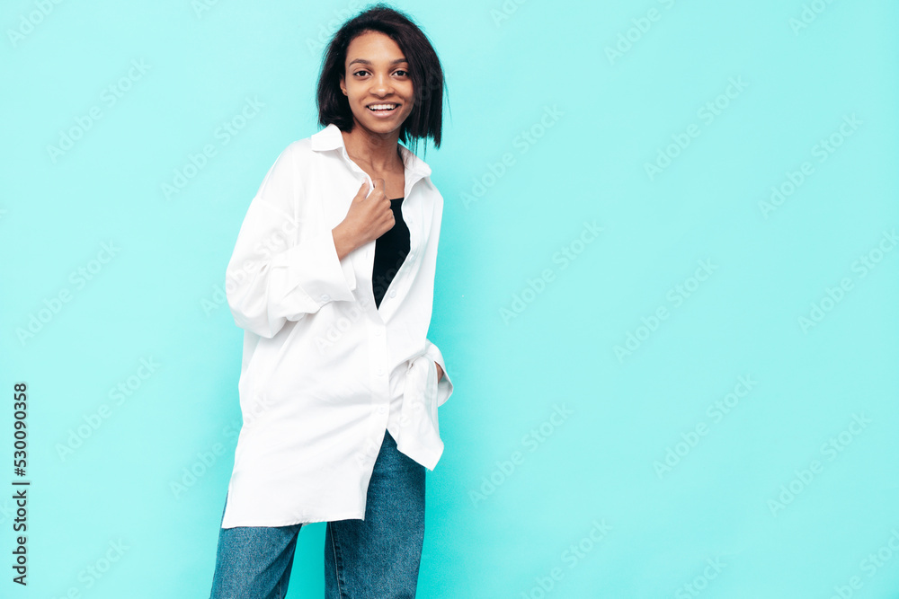 Portrait of young beautiful black woman. Smiling model dressed in summer jeans clothes. Sexy carefree female posing near blue wall in studio. Tanned and cheerful Portrait of young beautiful black woman. Smiling model dressed in summer jeans clothes. Sexy carefree female posing near blue wall in studio. Tanned and cheerful