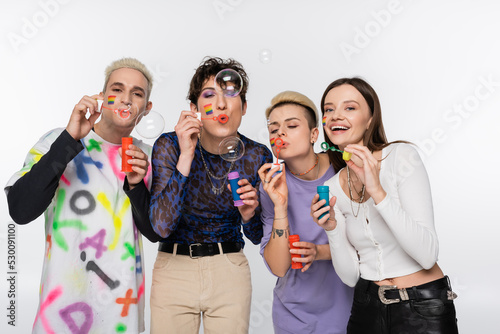 young people with lgbtq flags on faces blowing soap bubbles isolated on grey.