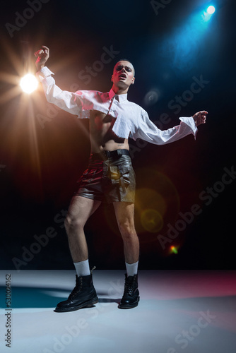Portrait of stylish man in makeup posing in short white shirt and leather shorts over black background in neon light. Expressive pose