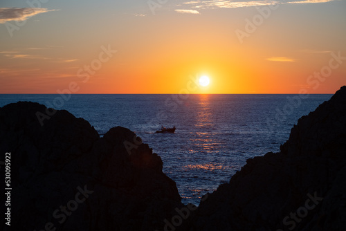 Colorful Capri sunset panorama near lighthouse “Faro“ at west end of famous italian island. Sun touching horizon and fisherboat passing. Silhouettes of cliff rocks forming a triangular shape.
