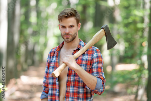 Lumbersexual man in lumberjack shirt holding axe on shoulder forest background photo