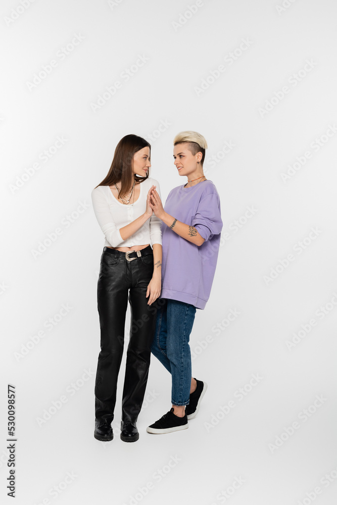full length of stylish lesbian women joining hands and looking at each other on grey background. full length of stylish lesbian women joining hands and looking at each other on grey background.