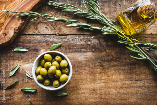 aceitunas variedad manzanilla en un bol, con una rama de olivo, una tabla de cortar y una aceitera de vidrio con aceite de oliva virgen,  sobre una mesa de madera rústica con espacio en blanco photo