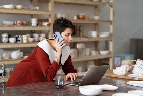 Young creative woman artisan artist ordering materials for ceramic work by phone while working in studio or workshop, female pottery shop owner talking with client via cellphone and typing on laptop © DimaBerlin