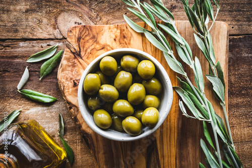 vista superior de un bol de aceitunas, con una rama de olivo y un frasco de vidrio con aceite de oliva, sobre una tabla de cortar de madera de olivo