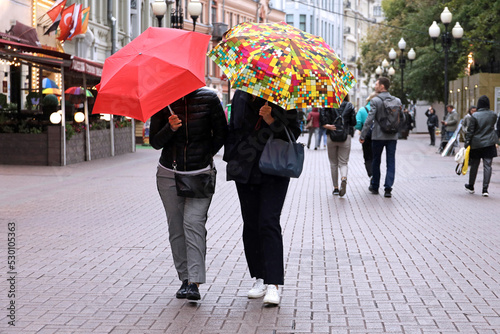 Rain in a city, two woman with umbrellas walk on a street on people background. Rainy weather in autumn