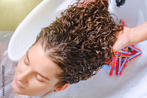 Professional hairstylist washes client’s hair with conditioner shampoo for washing after curling hair. Close up of curlers in hair. Creating hairstyles by a hairdresser in the salon. Shallow depth