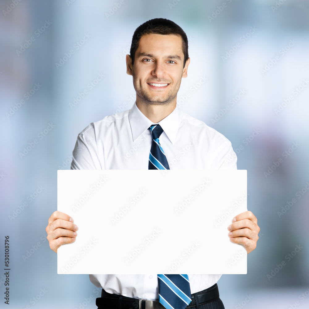 Happy smiling businessman in white shirt and tie, showing blank signboard with copy space area for text, over blurred office background. Confident business man holding paper board. Square composition. Happy smiling businessman in white shirt and tie, showing blank signboard with copy space area for text, over blurred office background. Confident business man holding paper board. Square composition.