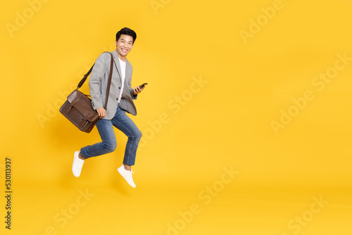 Attractive young Asian businessman jumping and typing smartphone isolated on yellow background