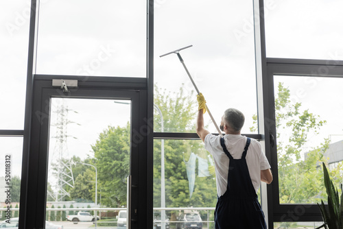 back view of man in overalls washing large office windows with window squeegee.