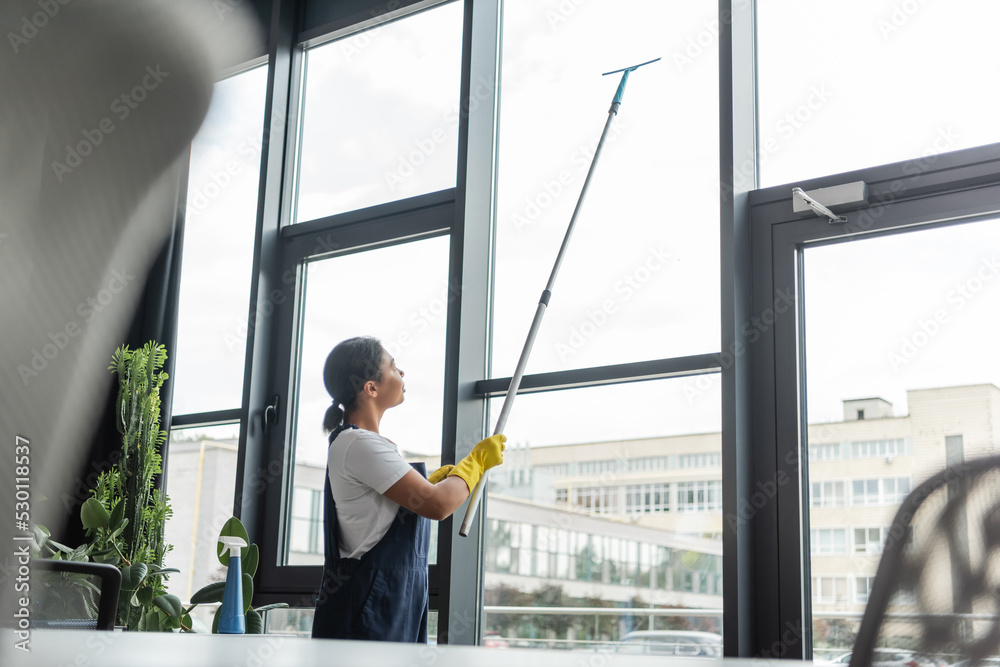 bi-racial woman in workwear washing large office windows on blurred foreground. bi-racial woman in workwear washing large office windows on blurred foreground.