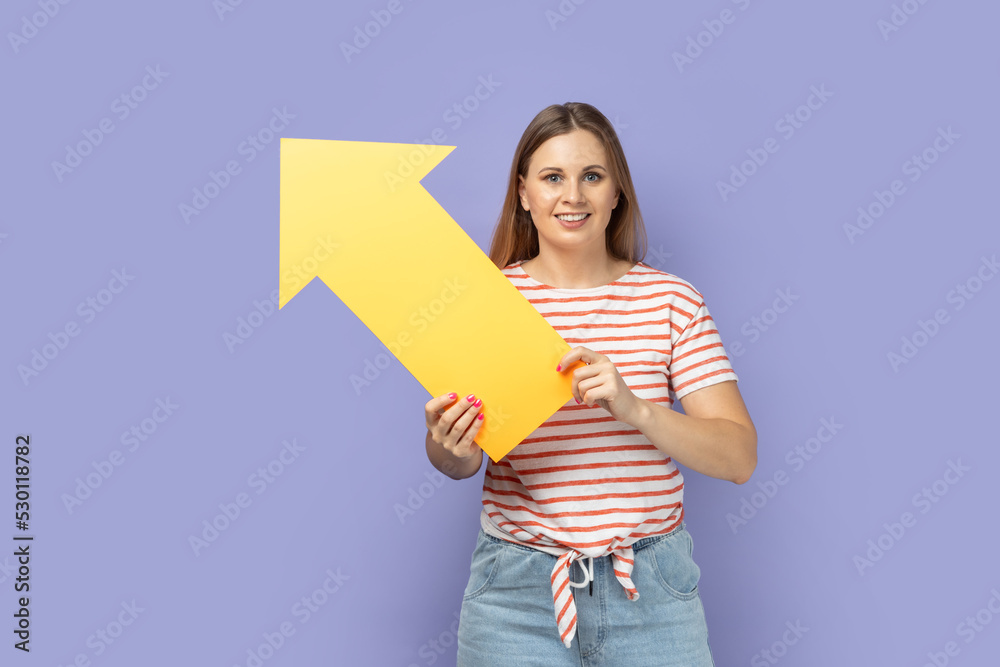 Portrait of delighted cute blond woman wearing striped T-shirt holding big yellow arrow indicating aside, looking at camera with smile. Indoor studio shot isolated on purple background. Portrait of delighted cute blond woman wearing striped T-shirt holding big yellow arrow indicating aside, looking at camera with smile. Indoor studio shot isolated on purple background.