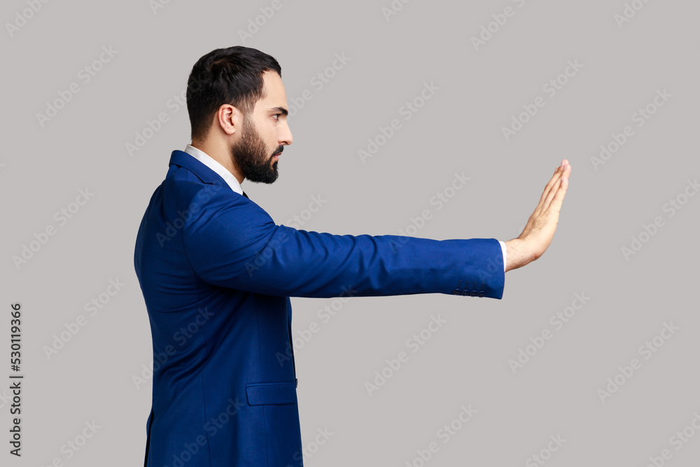 Side view of serious bearded man making stop gesture showing palm of hand, conflict prohibition warning about danger, stop bullying, wearing suit. Indoor studio shot isolated on gray background. Side view of serious bearded man making stop gesture showing palm of hand, conflict prohibition warning about danger, stop bullying, wearing suit. Indoor studio shot isolated on gray background.