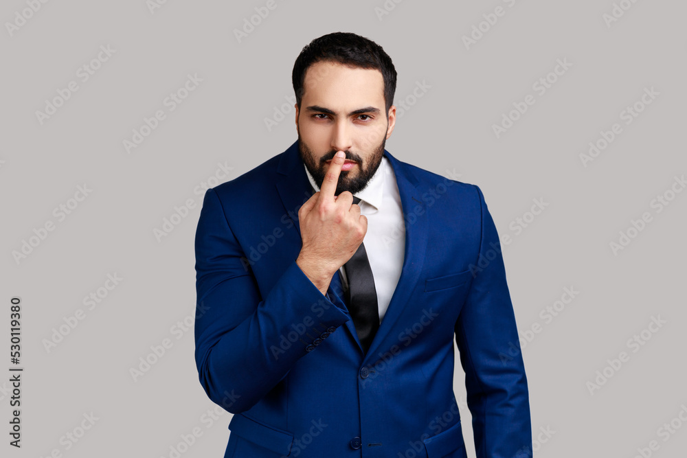 Bearded man looking with incredulous suspicious gaze and touching nose, gesturing you are liar, suspecting falsehood, wearing official style suit. Indoor studio shot isolated on gray background. Bearded man looking with incredulous suspicious gaze and touching nose, gesturing you are liar, suspecting falsehood, wearing official style suit. Indoor studio shot isolated on gray background.