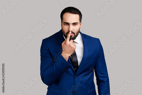 Bearded man looking with incredulous suspicious gaze and touching nose, gesturing you are liar, suspecting falsehood, wearing official style suit. Indoor studio shot isolated on gray background.