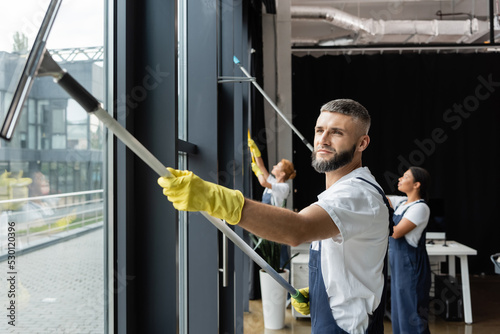 Canvastavla bearded man in rubber gloves washing windows near interracial team in office