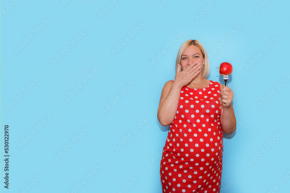 pregnant woman in a red polka dot dress with an apple on a fork in her hand closes her mouth with the other hand, on a blue background with copyspace, the concept of nutrition for pregnant women pregnant woman in a red polka dot dress with an apple on a fork in her hand closes her mouth with the other hand, on a blue background with copyspace, the concept of nutrition for pregnant women