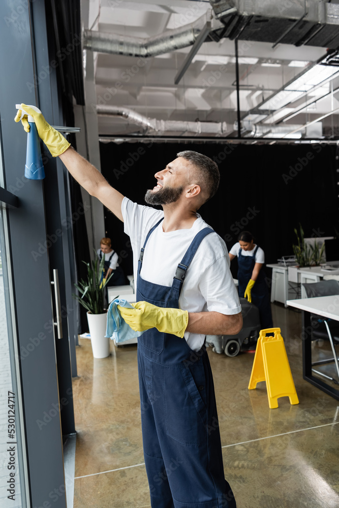 custom made wallpaper toronto digitalbearded man in workwear cleaning window with detergent near multiethnic women working in office.