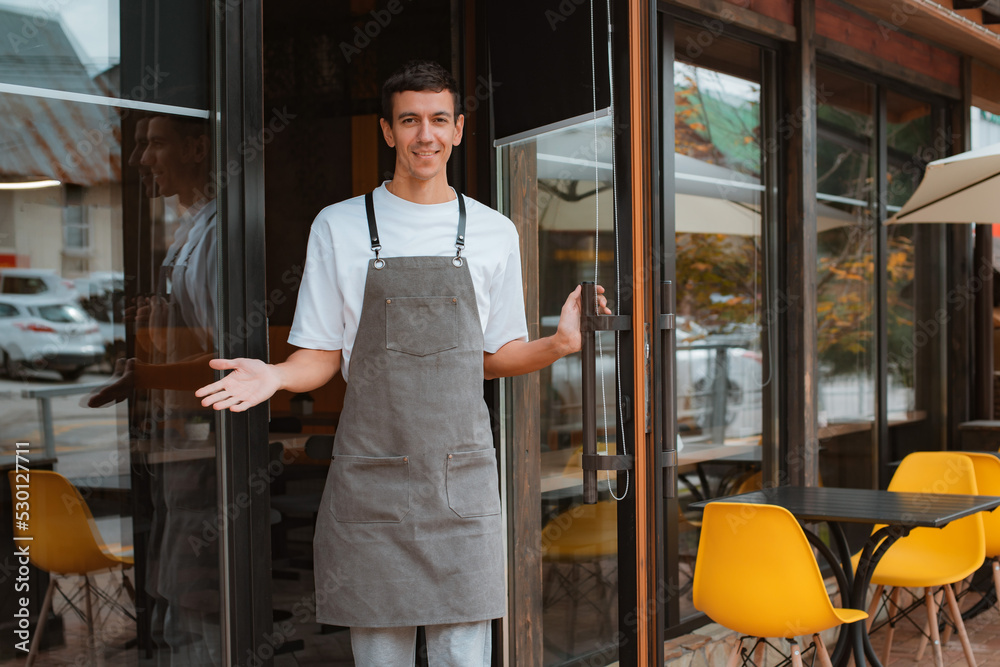 barista or waiter cafe or coffee shop owner against entrance, gesture inviting you to visit, smiling guy in apron standing outdoors being proud of his small local business