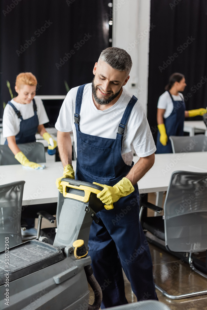 smiling bearded man with floor scrubber machine working near interracial women on blurred background. smiling bearded man with floor scrubber machine working near interracial women on blurred background.
