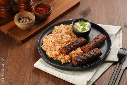 Traditional south european skinless sausages cevapcici made of ground meat and spices on black plate on dark wooden board, vegan substitute made of soy protein, with rice and sour cream