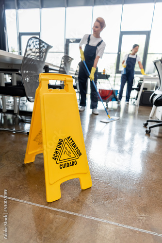 selective focus of warning sign board near blurred interracial women cleaning office.