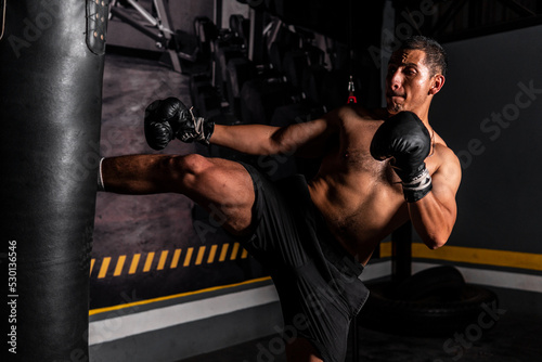 High quality photography. Shirtless martial artist training with a boxing bag in a gym with a dark background. Latino man kicking a boxing bag training fighting sport.