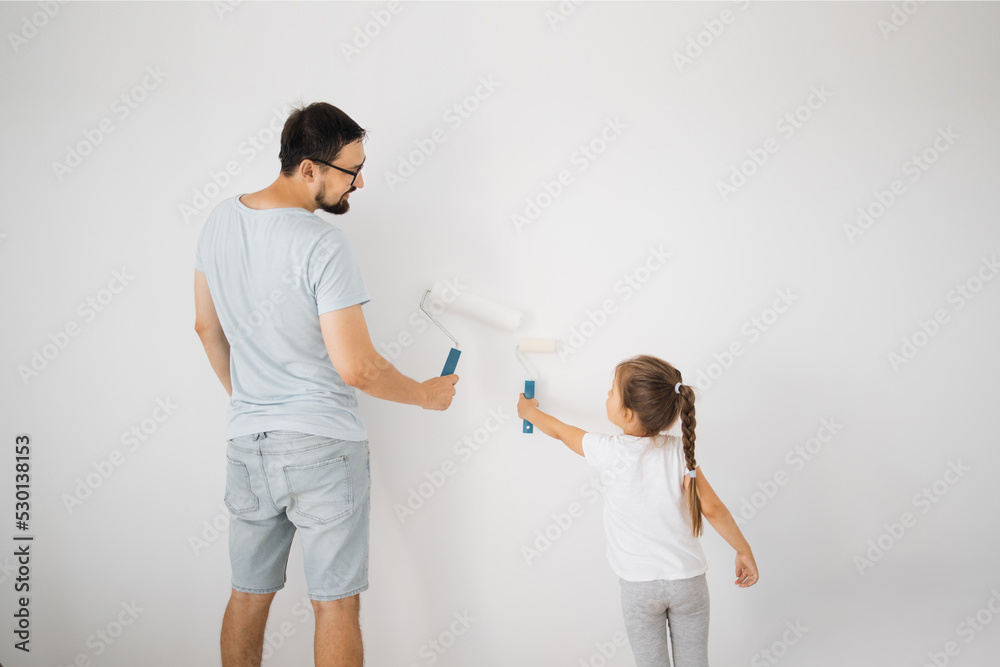 Fototapeta premium Repair in the apartment. Back view of happy family of father and little cute child daughter paints the wall with white paint.