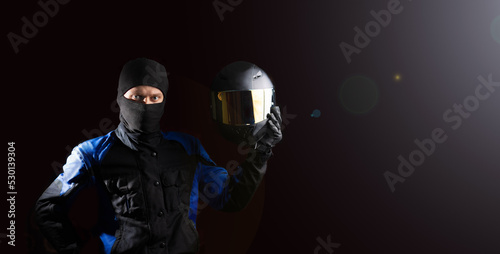 Fototapeta Naklejka Na Ścianę i Meble -  male motorcyclist in a motorcycle outfit and a balaclava puts on a helmet on a dark background. The concept of protective equipment for a biker.