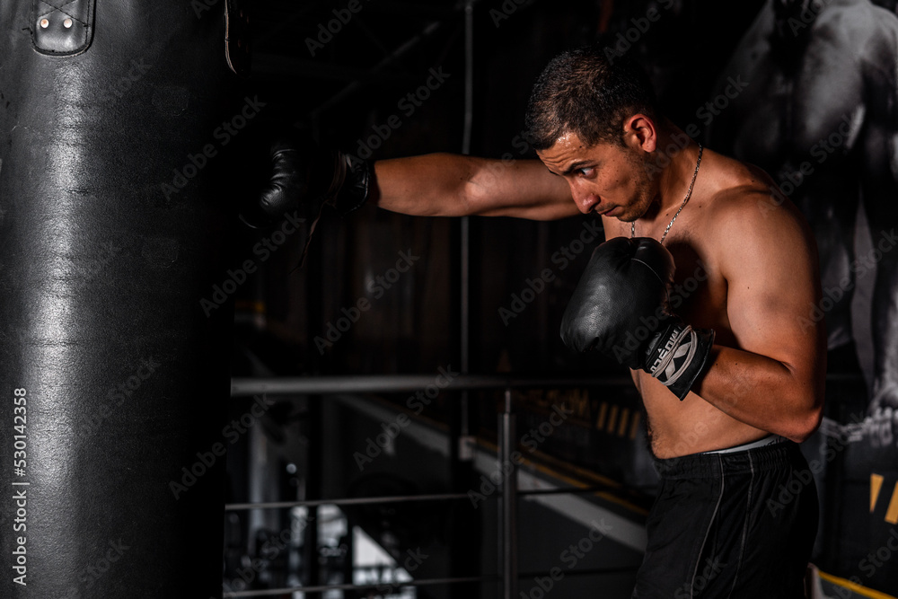 High quality photography. Shirtless, muscular Latino man throwing a punch at a boxing bag with gloves on. Latin man training in a boxing gym.