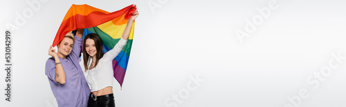 young and joyful lesbians with lgbtq flag looking at camera isolated on grey, banner.