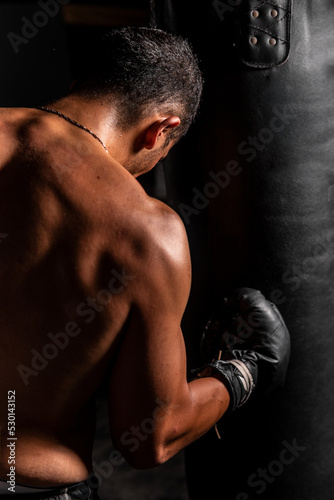 High quality photography. Back of a muscular Hispanic man hitting a heavy bag. Back of a man boxing with great force. © Wacha Studio