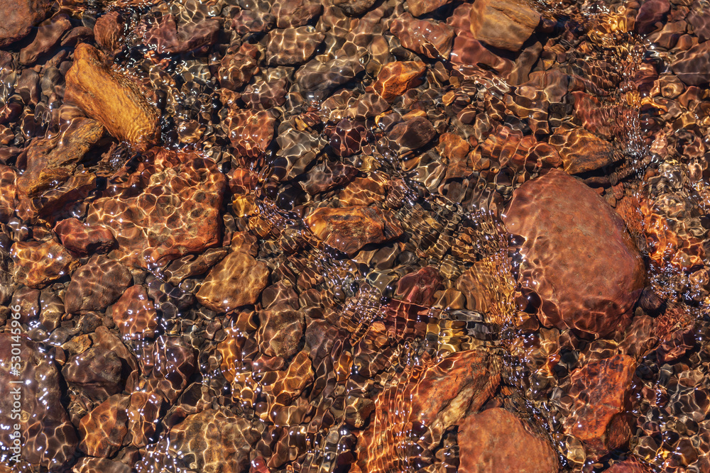 Nature background of clear water stream with motley stony bottom in sunlight. Colorful mountain stream texture with mineral water in bright sun. Pattern of sunlit multicolor stones in spring water.