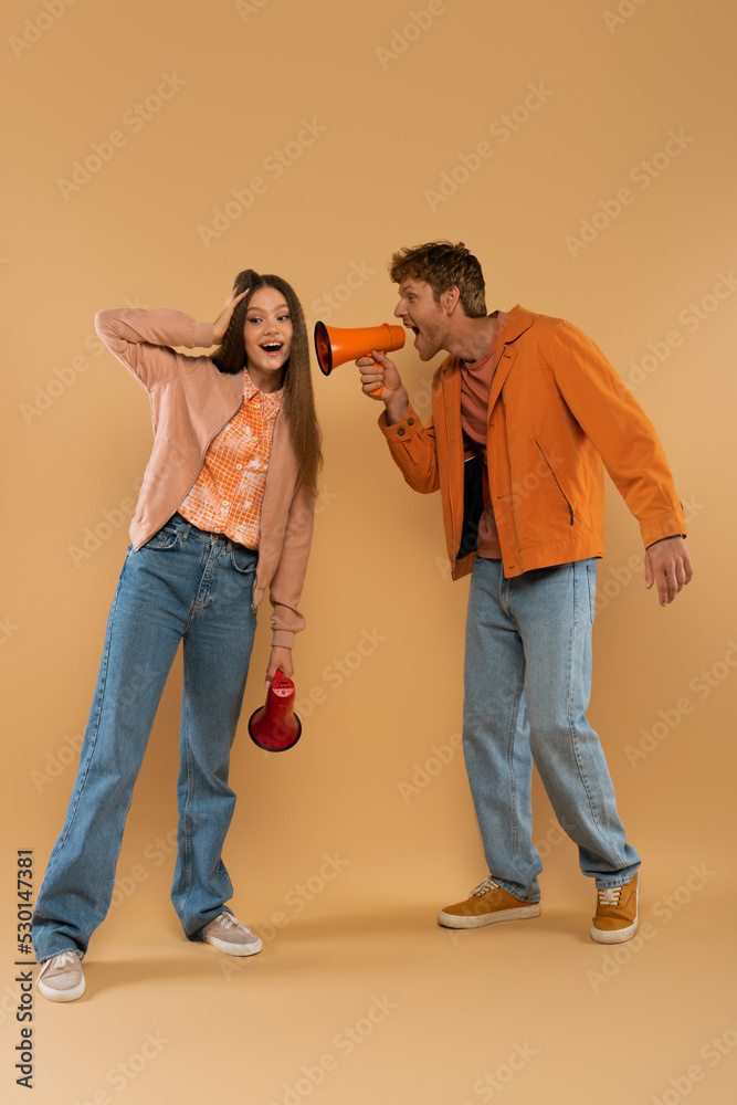 full length of redhead man screaming in loudspeaker near amazed girlfriend on beige.