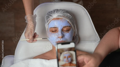 Female beauty business owner making promo photos of woman's client face with cosmetic skincare product on by smartphone for social medias and promotions. Beauty industry, spa and skincare concept.