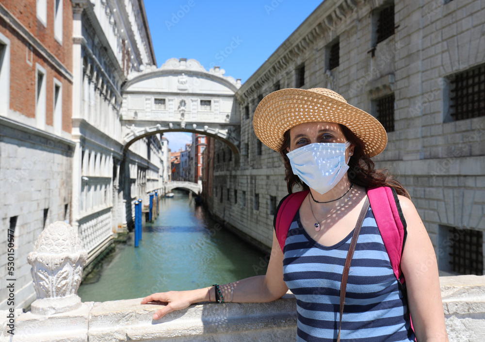 young female tourist with big straw hat surgical mask and in the background the very famous Bridge of Sighs in Venice in Italy