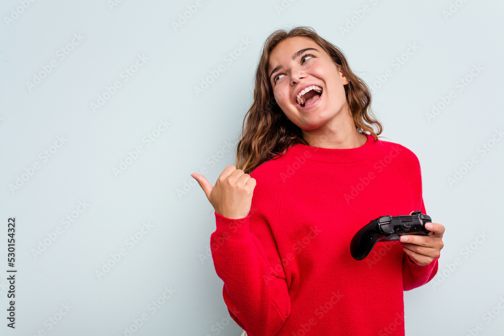 Naklejka premium Young caucasian gamer woman holding a game controller isolated on blue background points with thumb finger away, laughing and carefree.