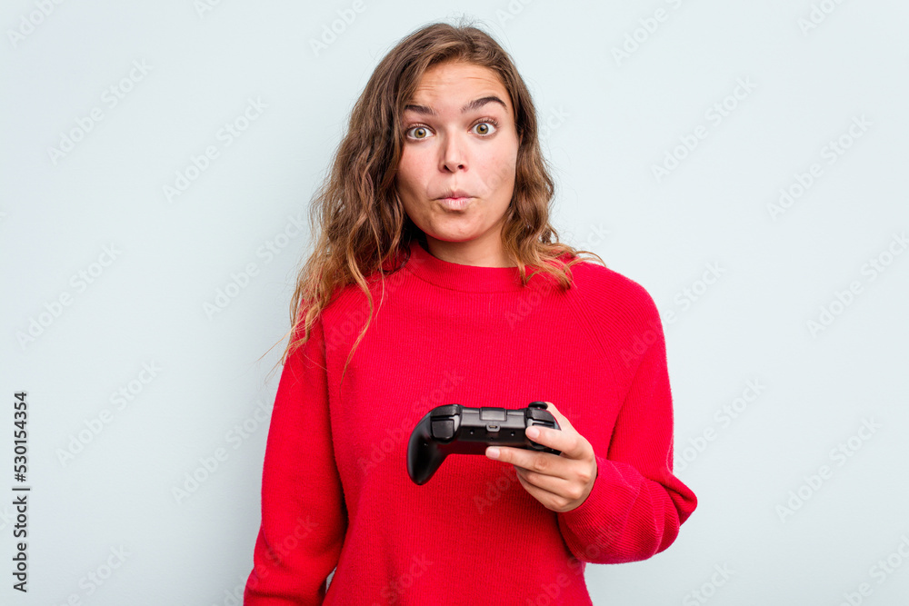 Young caucasian gamer woman holding a game controller isolated on blue background shrugs shoulders and open eyes confused. Young caucasian gamer woman holding a game controller isolated on blue background shrugs shoulders and open eyes confused.