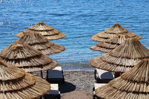 Fototapeta Naklejka Na Ścianę i Meble -  Empty pebble beach with wicker parasols and lounge chairs. Picturesque view to sea with blue water, summer resort