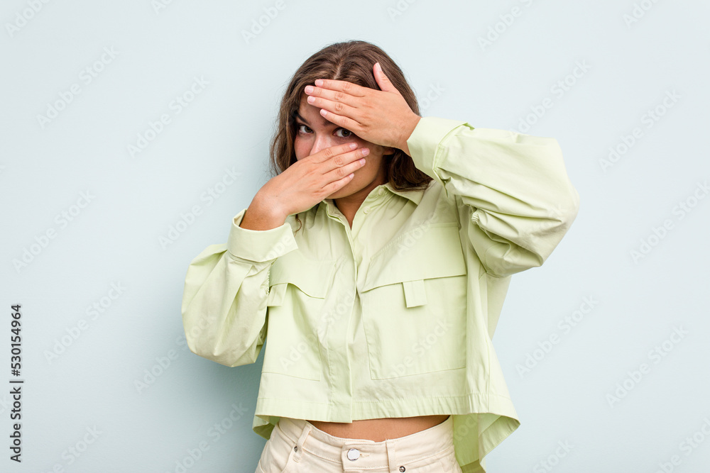 Young caucasian woman isolated on blue background blink at the camera through fingers, embarrassed covering face. Young caucasian woman isolated on blue background blink at the camera through fingers, embarrassed covering face.