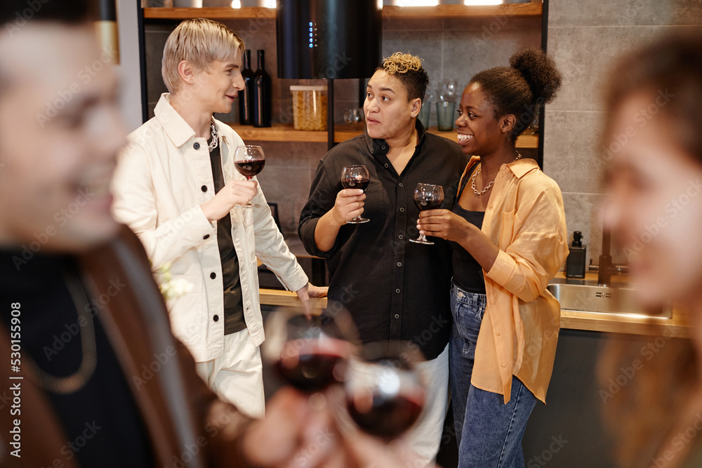 Obraz premium Group of young intercultural friends with glasses of red wine chatting to one another in the kitchen while enjoying home party
