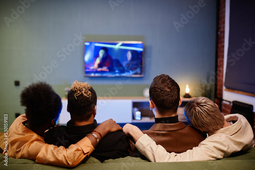 Rear view of two young affectionate homosexual couples watching movie while sitting on couch in front of tv set at leisure or weekend