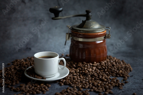 Hot coffee in a white coffee cup, a coffee grinder and a lot of coffee beans scattered around, on a dark background.