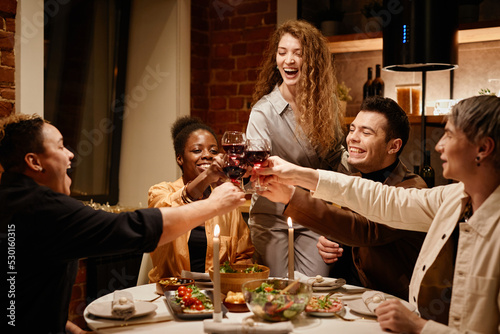 Young ecstatic friends clinking with red wine in wineglasses over dinner table served with homemade food and decorated with burning candles