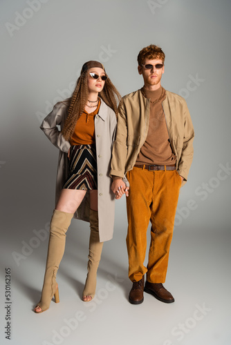 full length of young and stylish couple in autumnal outfits holding hands on grey.