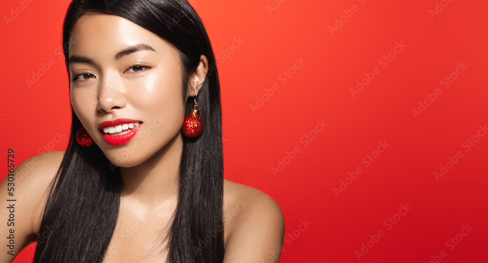 Fashion girl with red lips and beautiful smile, standing in christmas earrings and looking flirty at camera, coquettish smiling face. Asian woman posing against red background Fashion girl with red lips and beautiful smile, standing in christmas earrings and looking flirty at camera, coquettish smiling face. Asian woman posing against red background